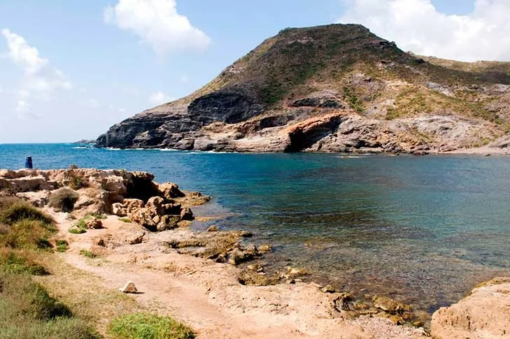 Cala Reona con el monte rocoso al fondo, el agua turquesa y un sendero de tierra entre matorral