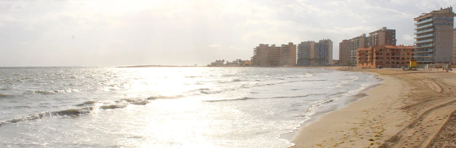 Orilla de Playa del Estacio al amanecer, con las olas del Mediterráneo y los edificios de La Manga al fondo