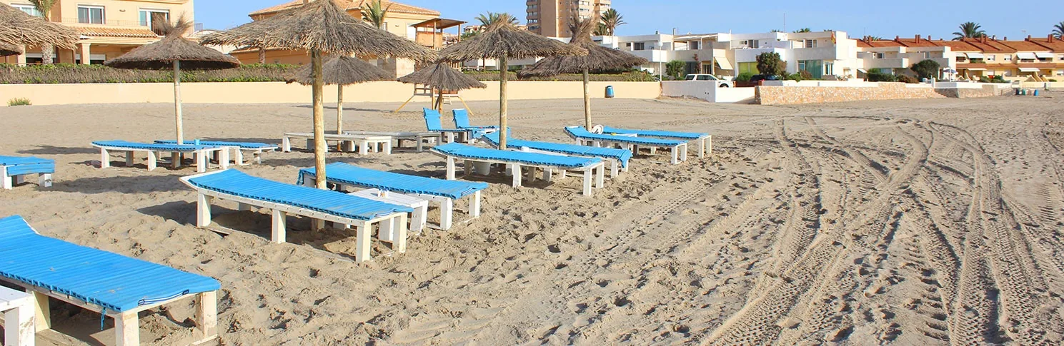 Hamacas azules alineadas bajo sombrillas de paja sobre la arena rastrillada de Playa Galúa