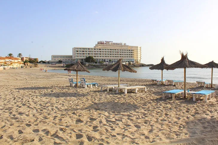 Sombrillas cónicas de paja y hamacas azules en Playa Galúa, con el hotel de La Manga al fondo