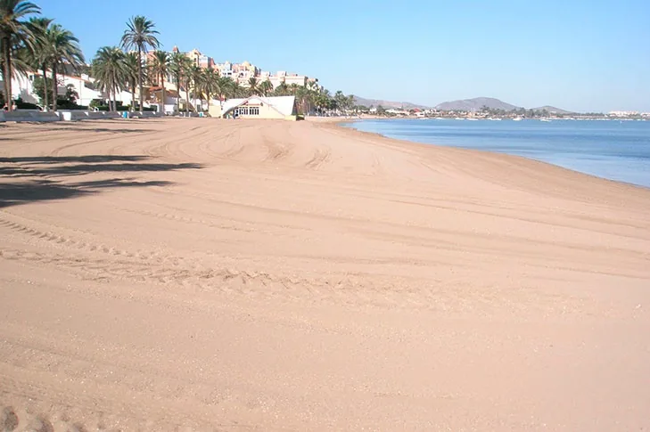 Arena rastrillada de Playa Honda con el paseo, edificios blancos y palmeras a un lado y el Mar Menor al otro
