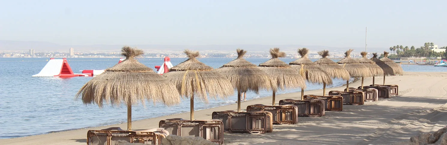 Sombrillas de paja y bancos de madera sobre la arena de Playa La Veneziola, junto al Mar Menor en calma