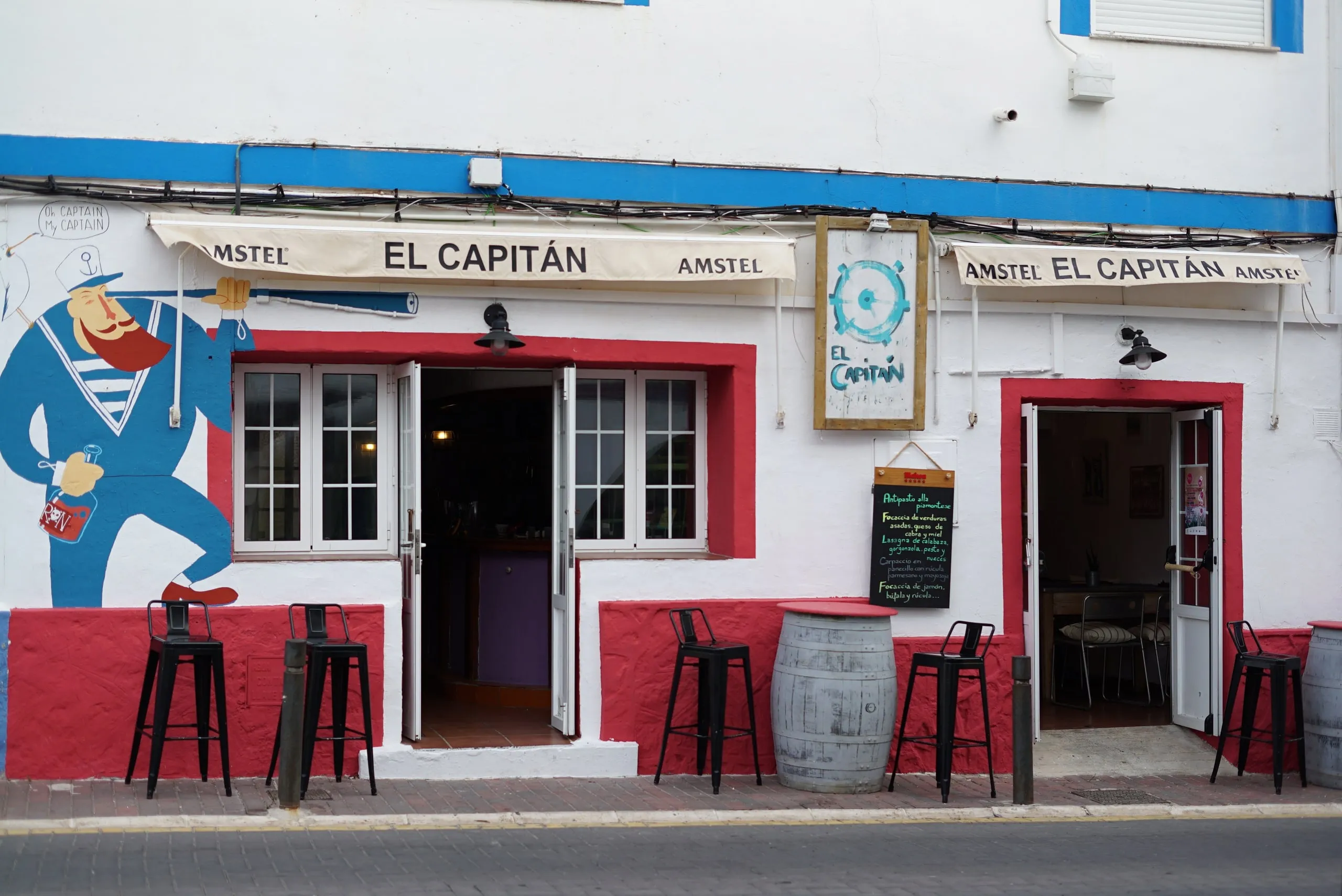 Fachada de El Capitán en Calle Marín, Cabo de Palos, con mural de capitán de barco y terraza con taburetes