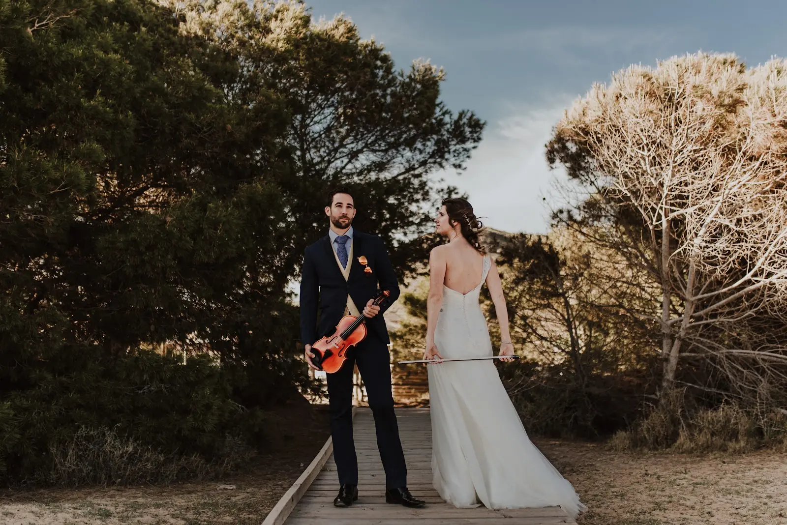 Novios posando con un violín en un camino de madera entre pinos, fotografía de José Espinosa